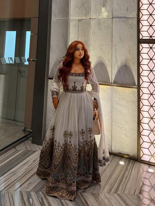 Woman in a long, patterned Gold Habesha Zuria standing in a modern indoor setting with geometric wall designs.