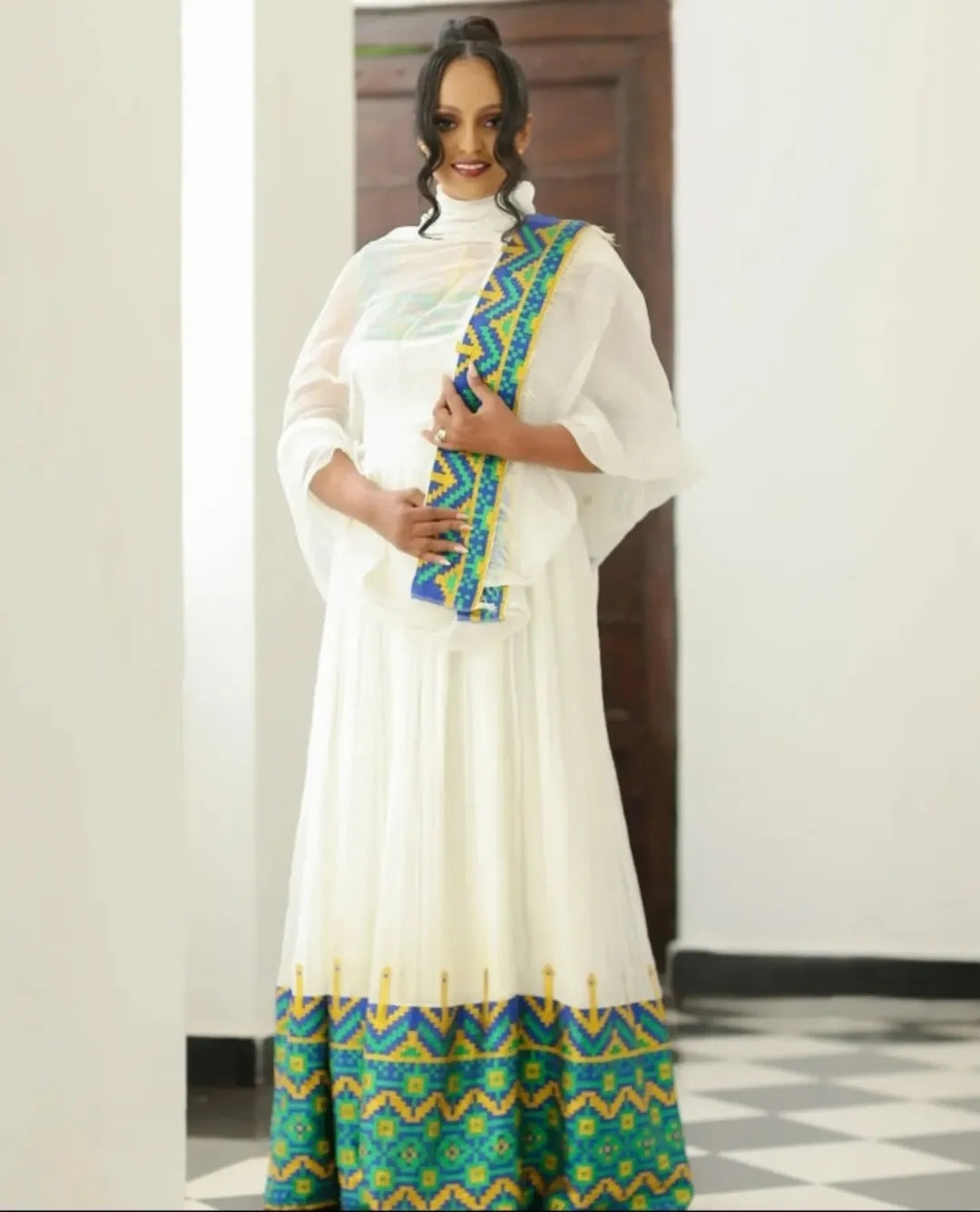 Woman wearing a white outfit with green and blue Ethiopian dress , standing indoors.