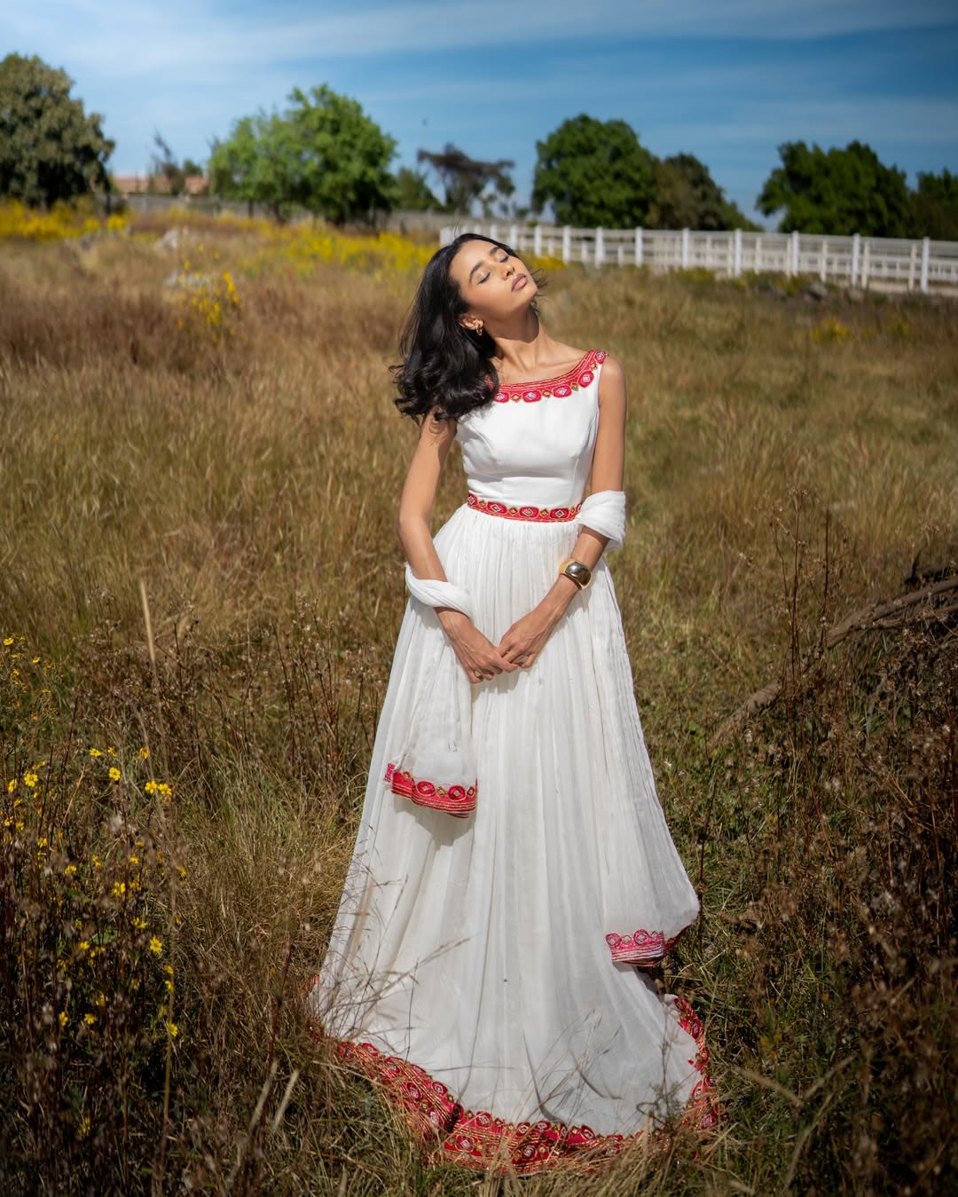 Woman in a habesha kemis with red accents standing in a field with trees and a fence in the background.