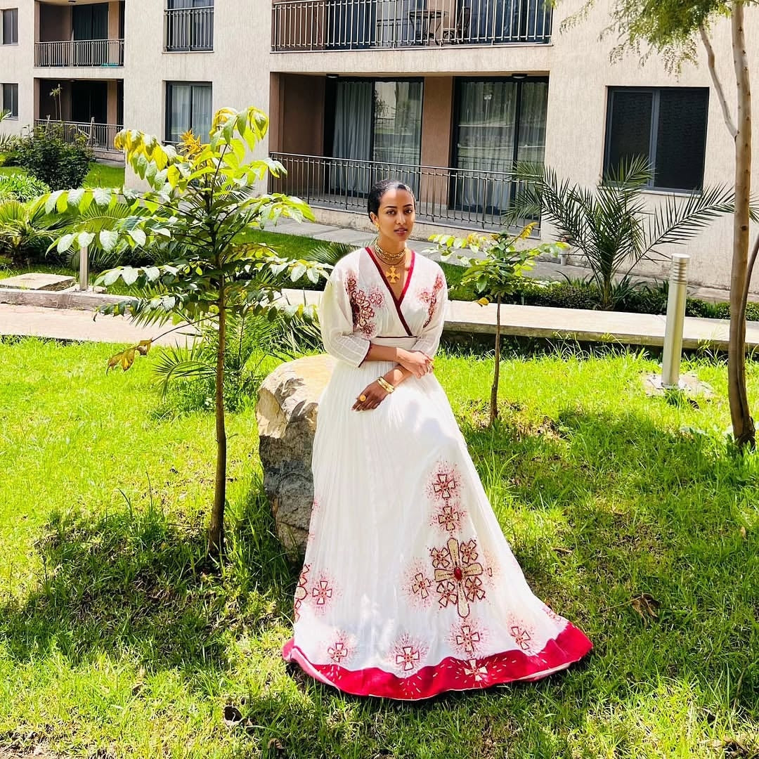 Woman in a white Habesha Zuria with red embroidery sitting on grass.