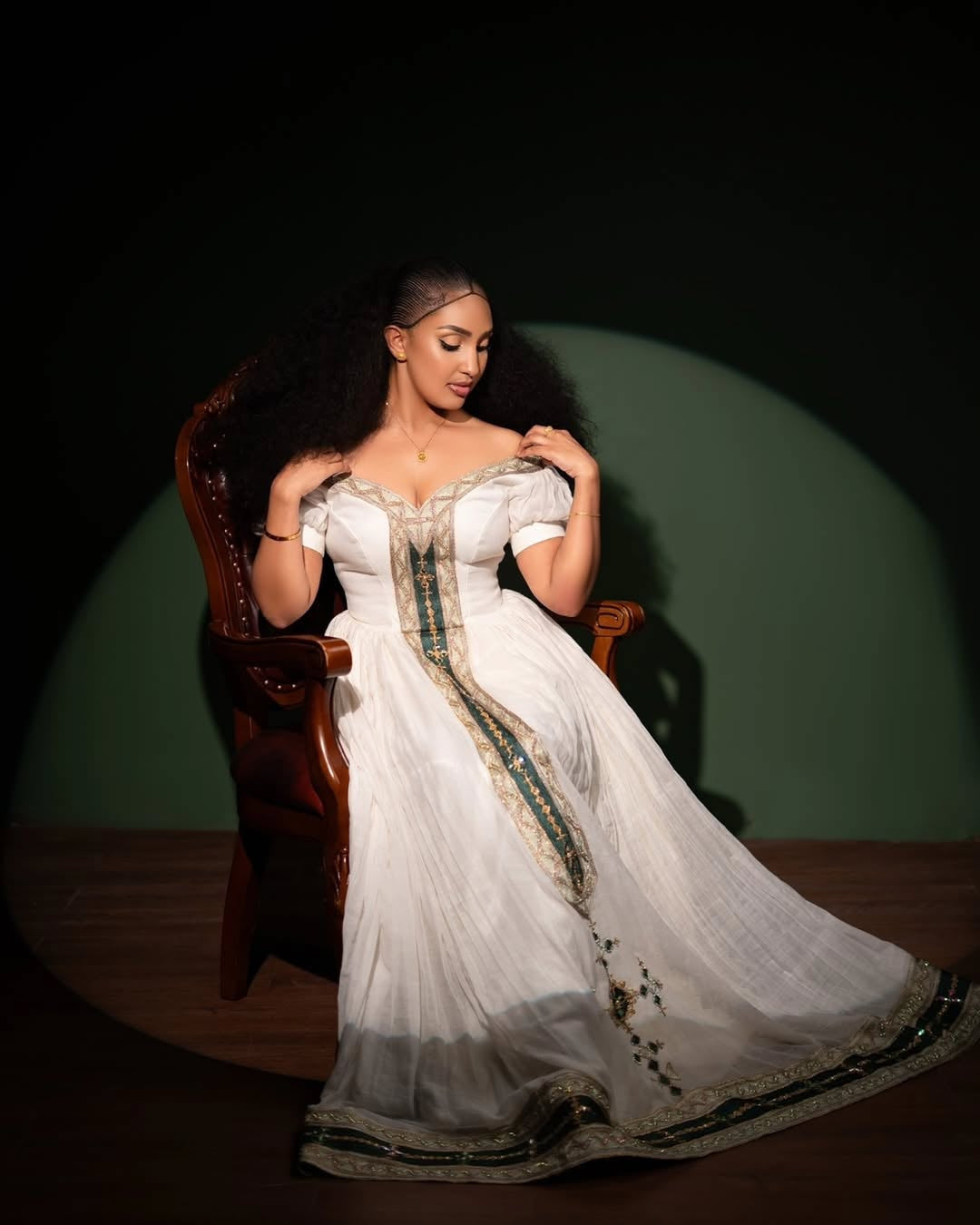 Woman in a white Ethiopian Dress with green patterns sitting on a wooden chair against a dark background