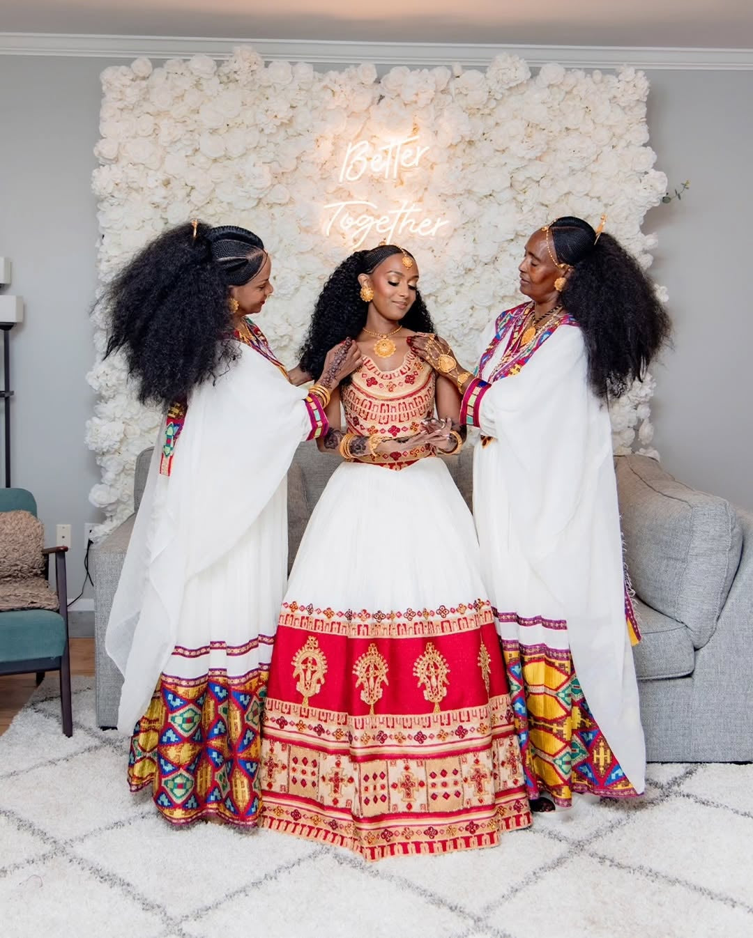 Three women in traditional Ethiopian dress standing together in a room with decorative floral wall.