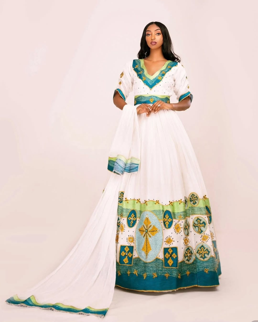 Woman wearing a white dress with green and blue Ethiopian dress patterns on a plain background