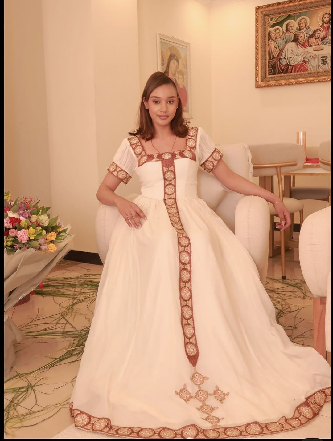 Woman in a white Ethiopian cultural dress with brown embroidery in a decorated room.