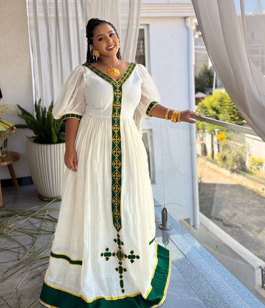 Woman wearing a white dress with green and gold details Ethiopian dress, standing in a home setting.