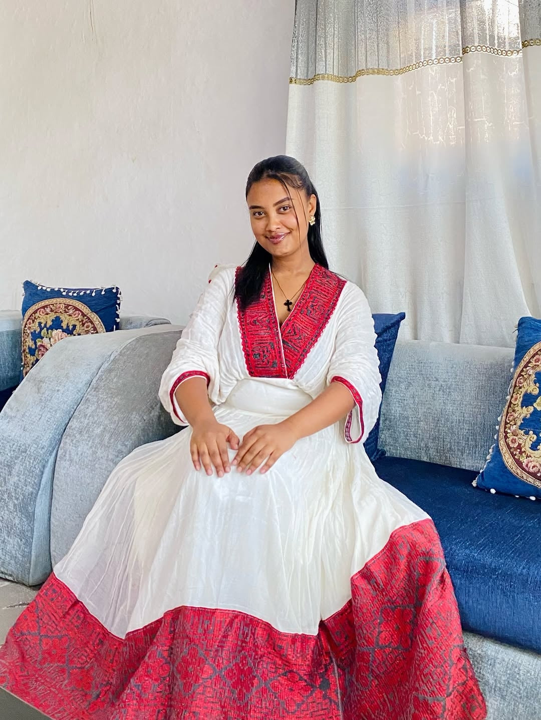 Woman in a white and red traditional Ethiopian dress sitting on a couch with decorative pillows.