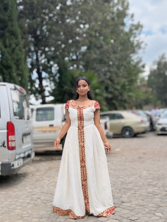 Woman in a white habesha dress with red patterns standing on a street with cars and trees in the background