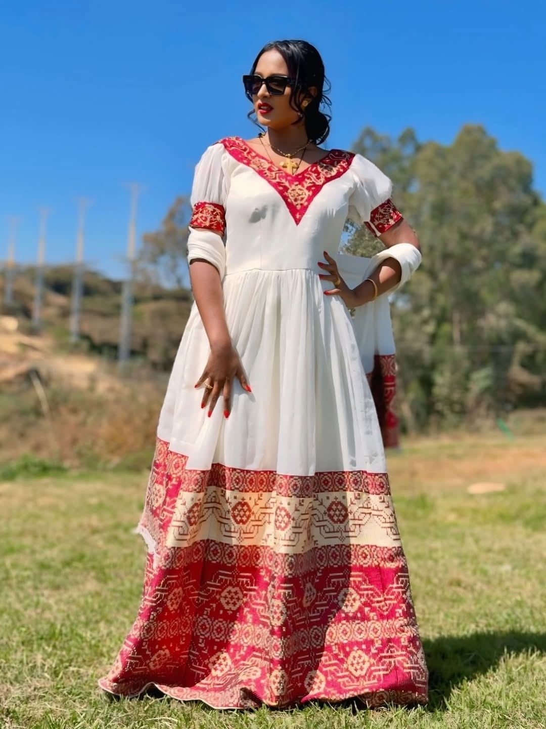 Woman wearing a white and red Ethiopian cultural dress with patterns standing outdoors on grass.