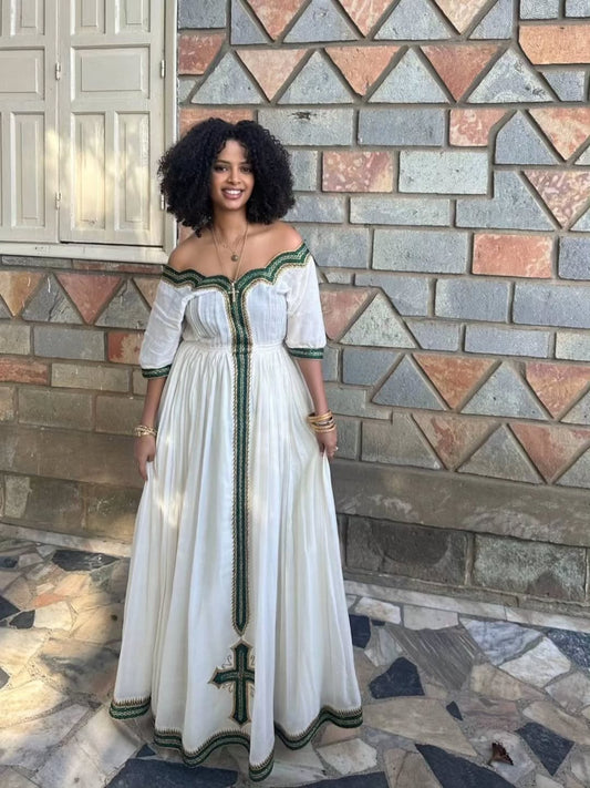 Woman in a white green Habesha Libs with green accents standing in front of a stone wall.