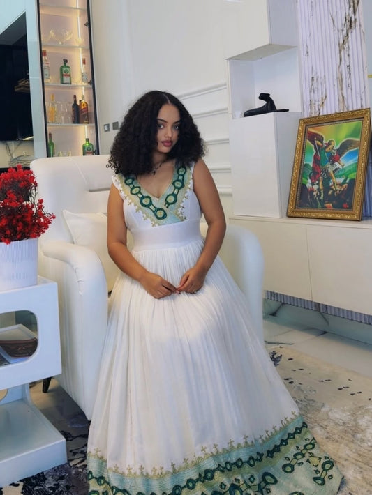 Woman in a white habesha dress with green embroidery sitting on a white chair in a modern living room.