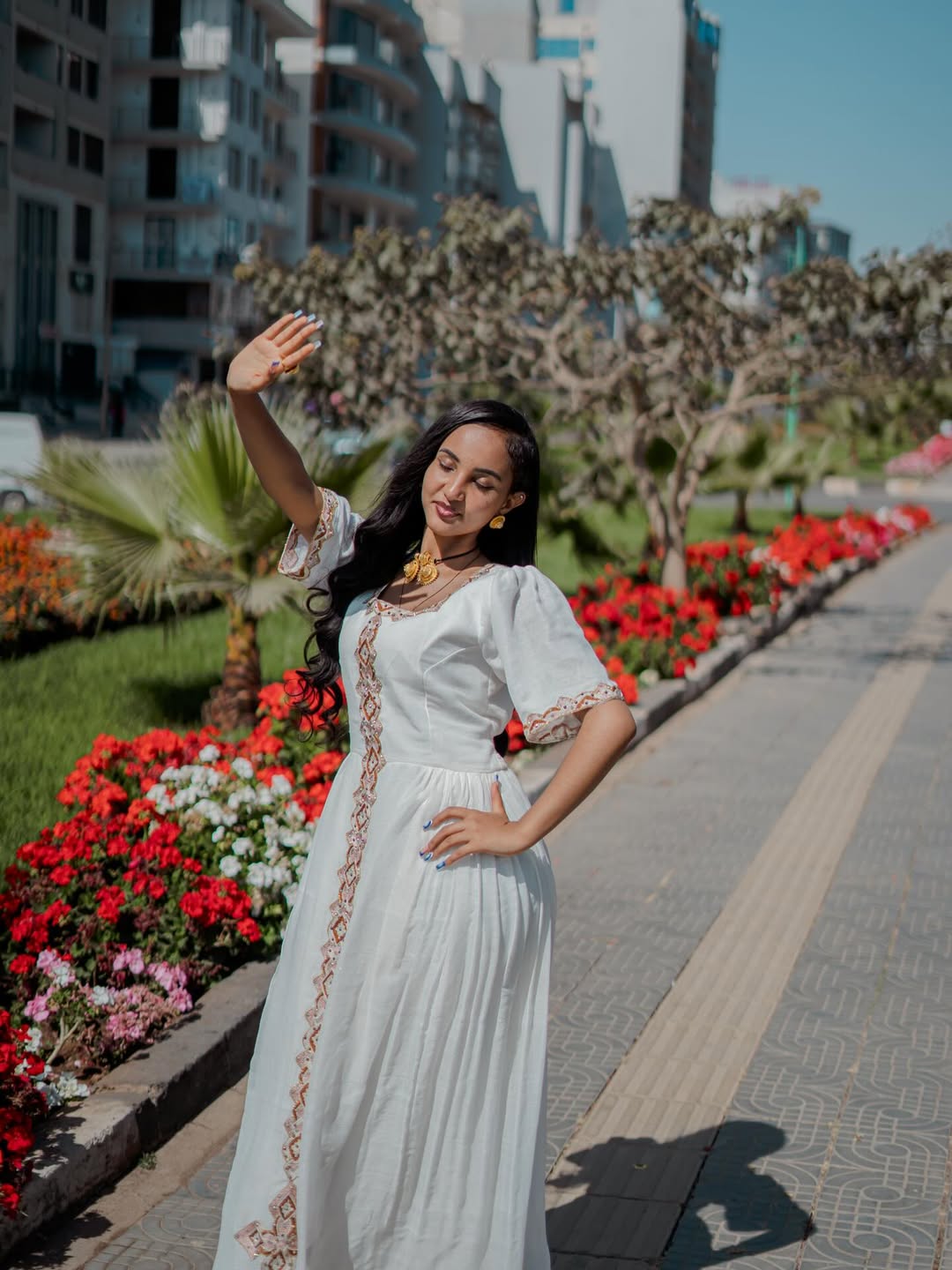 Woman in a habesha dress standing on a sidewalk with flowers and buildings in the background