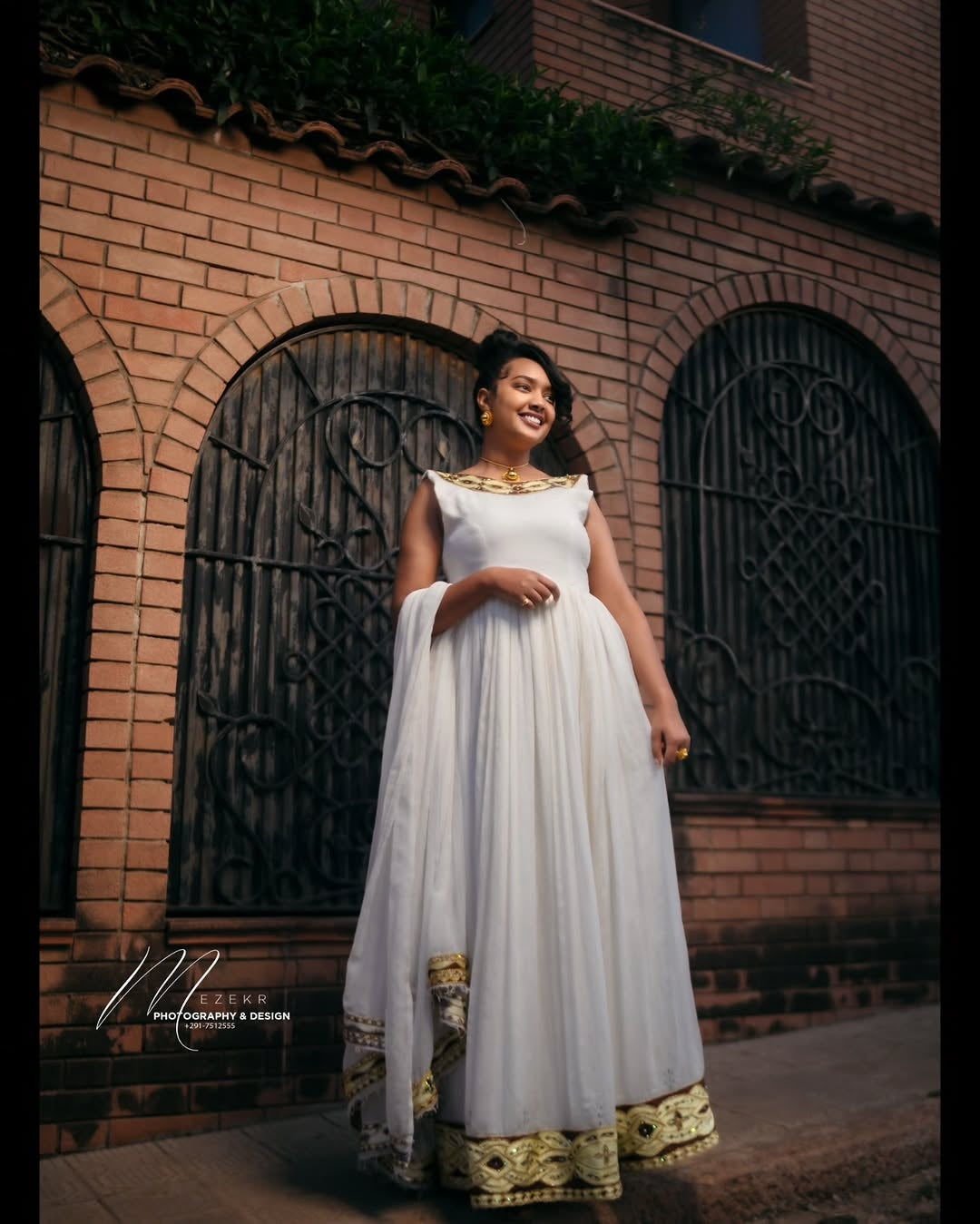 Woman in a white Habesha Zuria with gold details standing in front of a brick building.