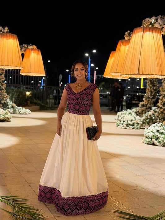 Woman in a Ethiopian Traditional Dress with a dark purple top and white skirt standing in an outdoor decorated area at night.