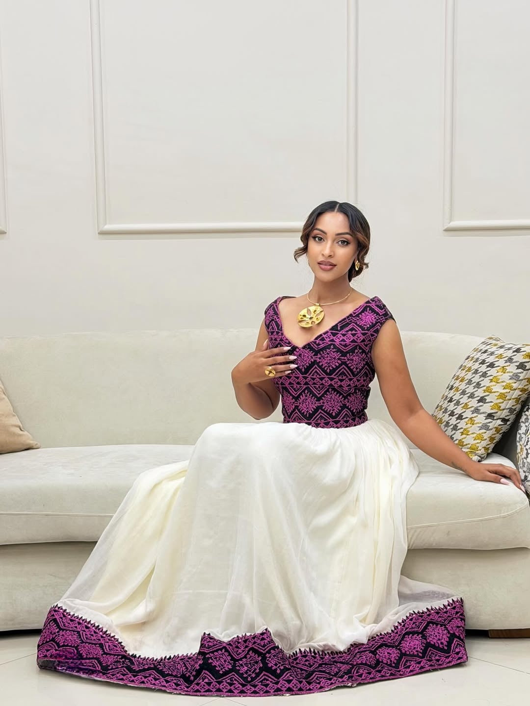 Woman in a purple and white Ethiopian Traditional Dress sitting on a couch.