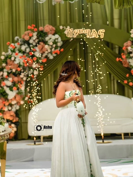 Woman in a white Habesha Bridal Dress holding a bouquet in front of a decorated stage with flowers and lights.