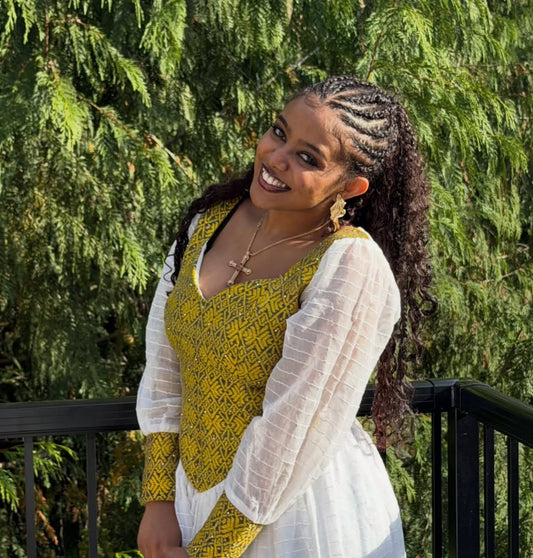 A smiling woman with Habesha Dress stands outdoors in front of green foliage, wearing a yellow and black patterned top with sheer white long sleeves and a white skirt.