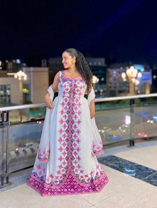 Woman in a white and pink patterned habesha kemis standing on a balcony at night.