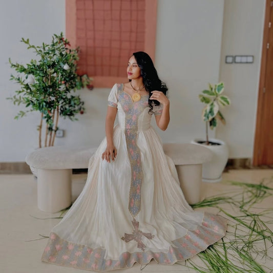 Woman in a white Ethiopian cultural dress with floral patterns standing in a room with plants and a bathtub.
