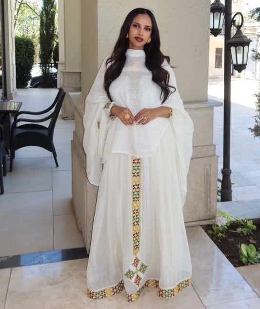 Woman in a white traditional Ethiopian cultural dress with colorful embroidery standing outdoors.