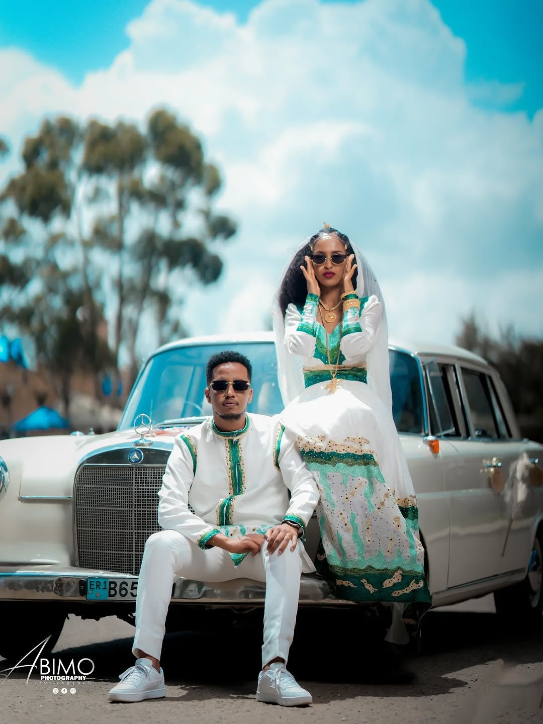 Two people in formal attire sitting on a vintage car with a scenic background