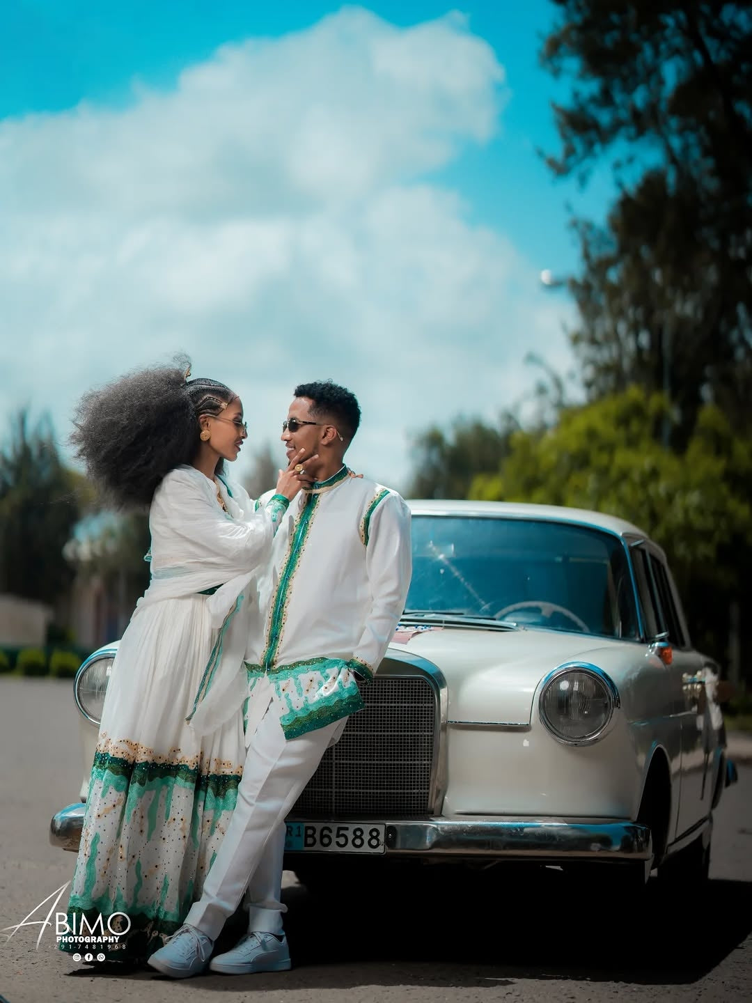 Couple in formal attire standing next to a vintage car with a scenic background