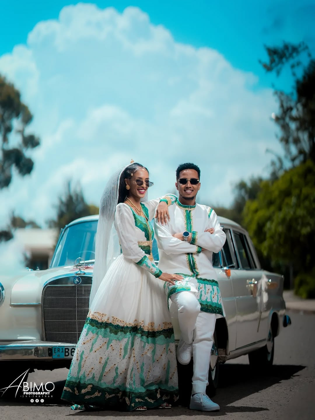 Couple in traditional attire standing in front of a vintage car with a clear blue sky.