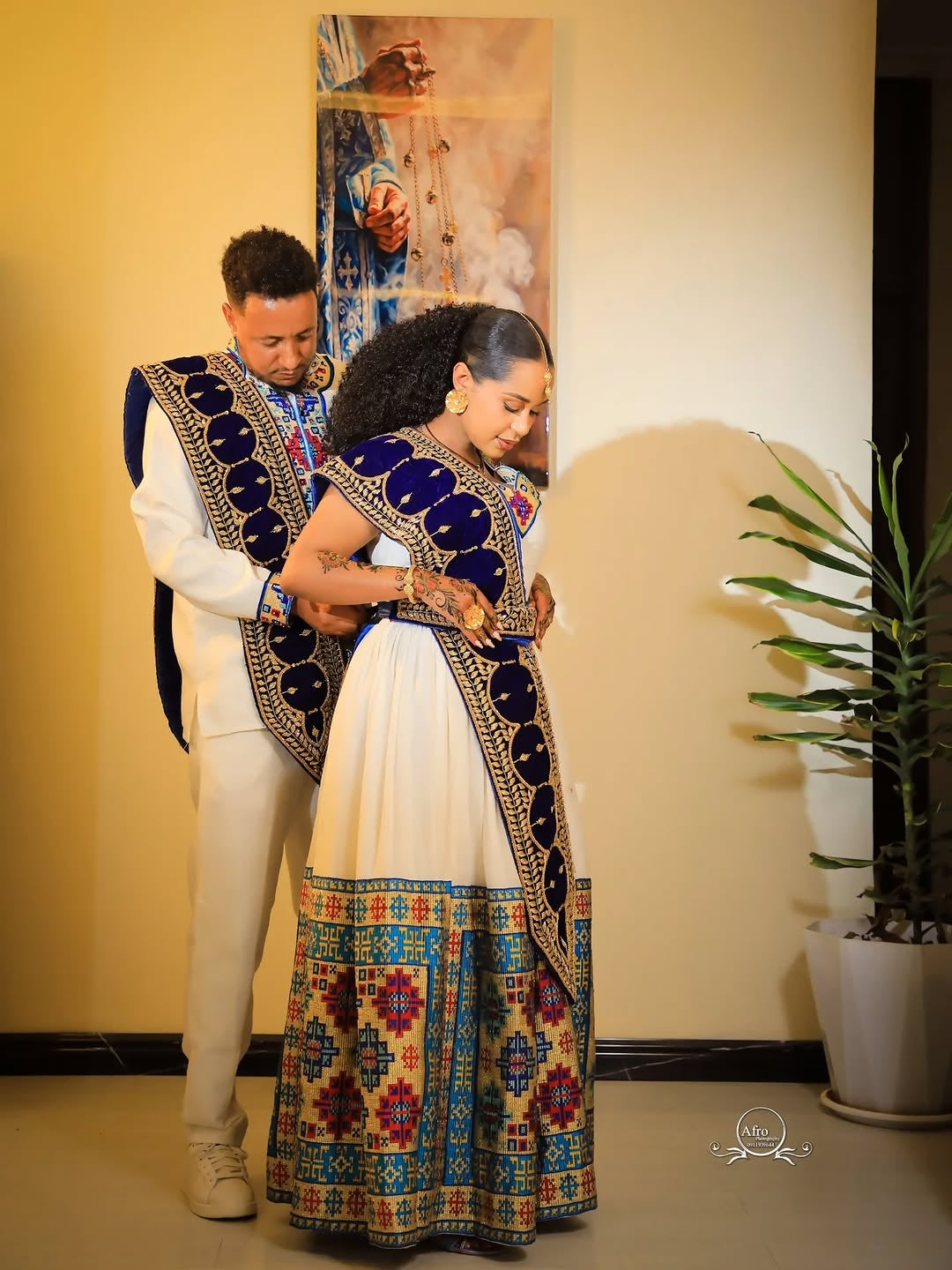 Couple in traditional attire standing indoors with a decorative wall and plant in the background
