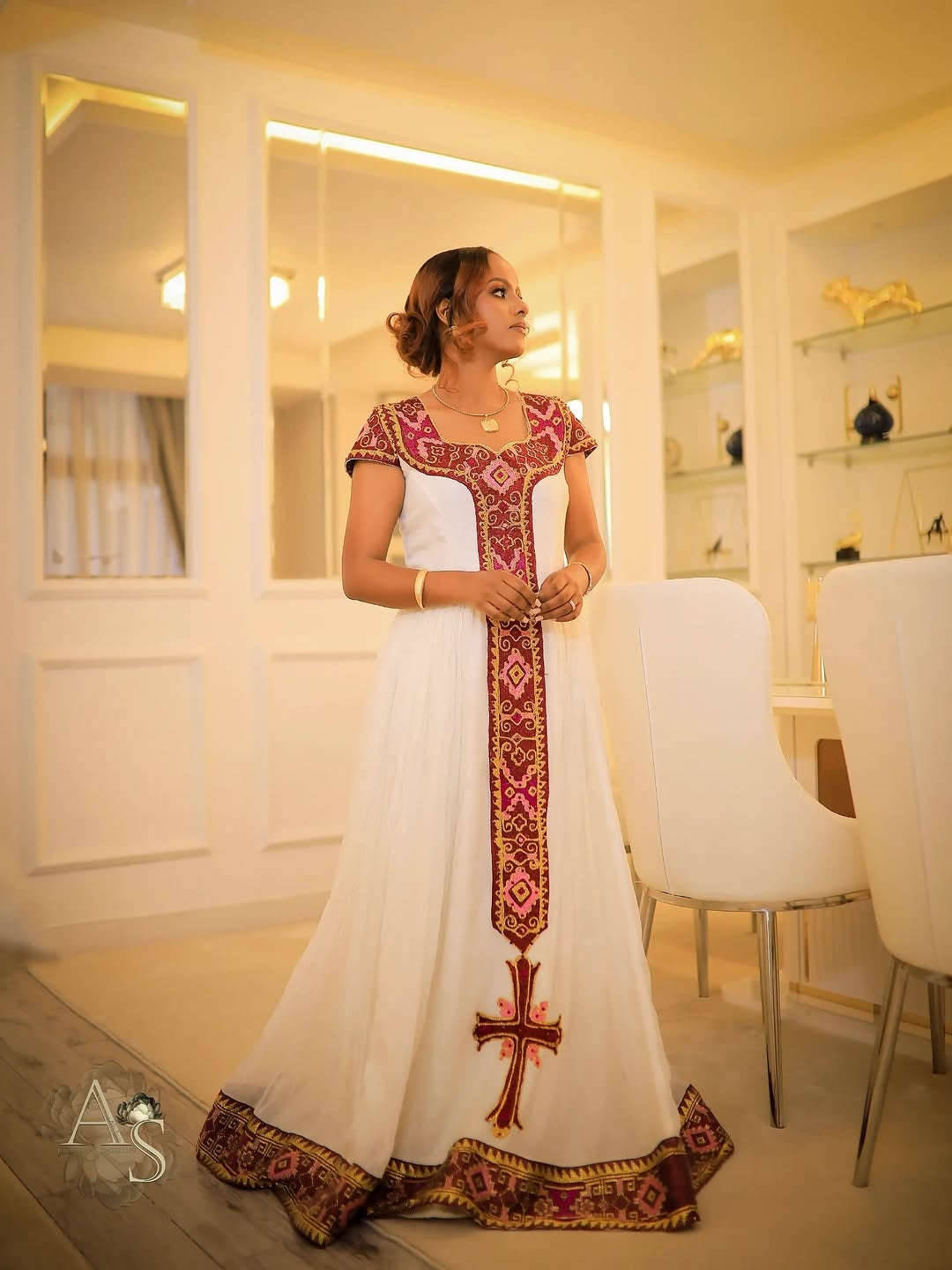 Woman in a white dress with red and gold habesha dress standing in a modern interior setting.