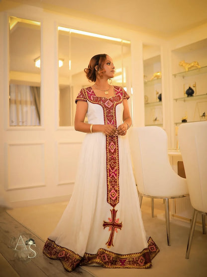 Woman in a white dress with red and gold habesha dress standing in a modern interior setting.