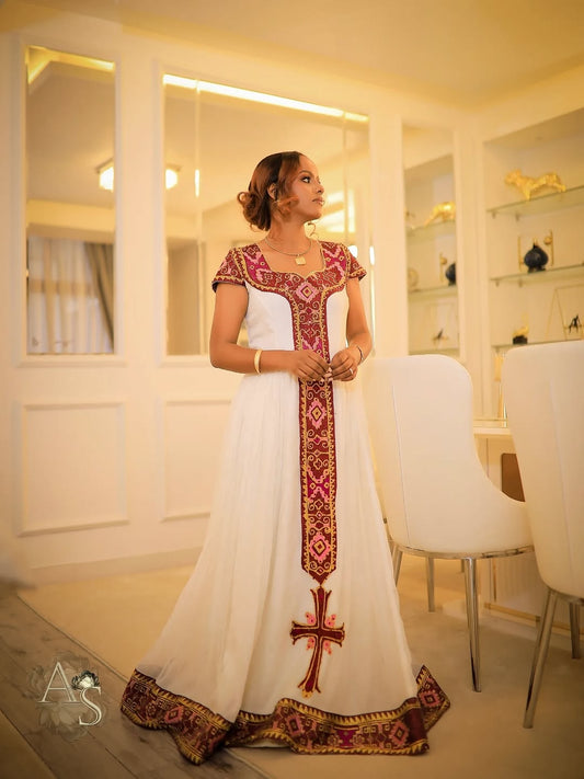 Woman in a white dress with red and gold habesha dress standing in a modern interior setting.