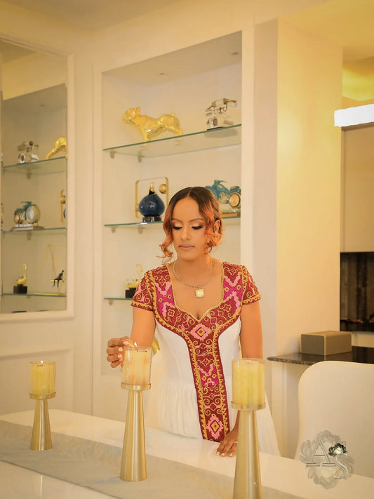 Woman in a decorative habesha dress holding candles in a room with shelves displaying decorative items.