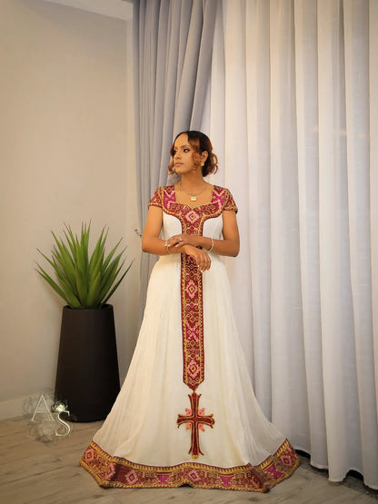 Woman wearing a white habesha dress with red and gold embroidery standing in a room with curtains and a plant.