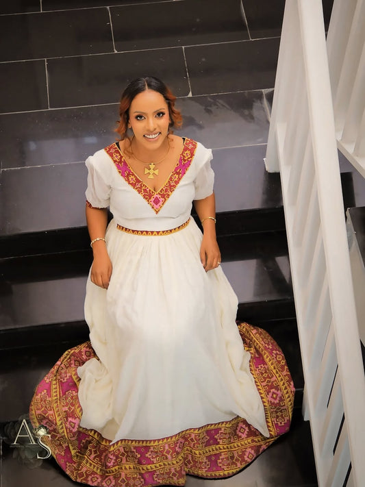 Woman in a white habesha dress with red and gold embroidery sitting on a staircase.