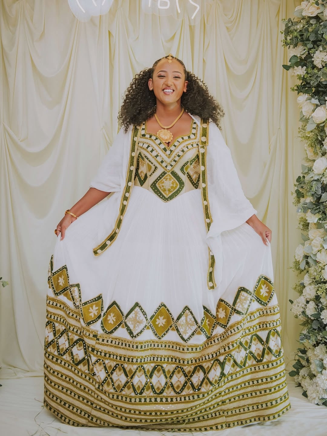 Woman wearing a white and gold traditional outfit with a decorative pattern, standing against a decorated backdrop.