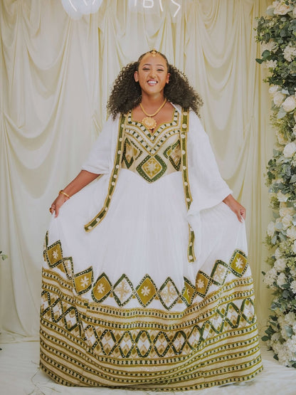 Woman wearing a white and gold traditional outfit with a decorative pattern, standing against a decorated backdrop.