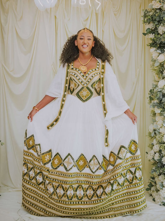 Woman wearing a white and gold traditional outfit with a decorative pattern, standing against a decorated backdrop.