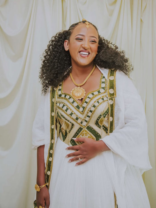Woman wearing a traditional outfit with intricate patterns and gold accents against a white curtain background.
