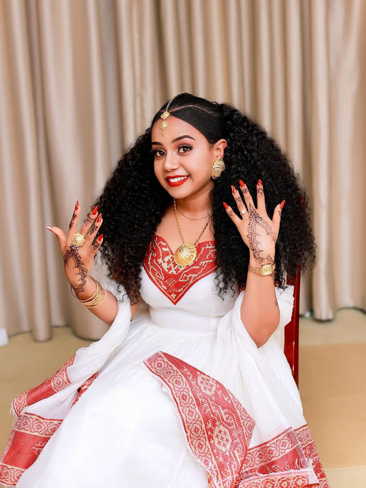 Woman in a white and Red Ethiopian Traditional Dress with jewelry and henna on her hands, sitting against a beige curtain.