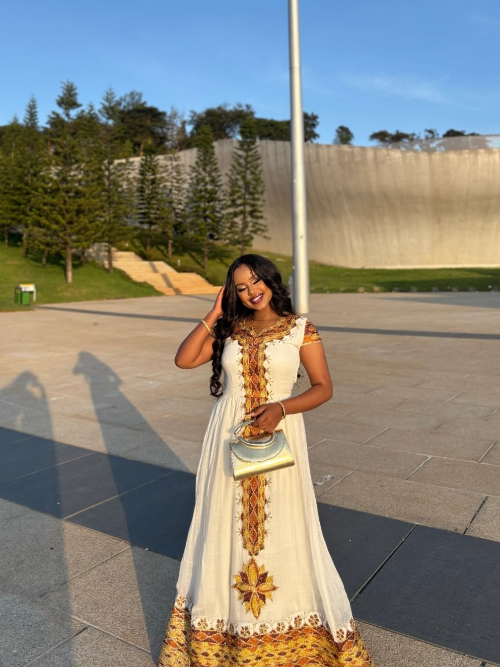 A woman wearing a long white Ethiopian clothing with gold and orange embroidery stands outdoors in a paved plaza with trees and a concrete wall in the background, holding a small metallic clutch.