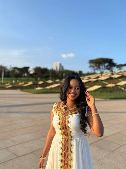 A woman wearing a white sleeveless Ethiopian clothing with a gold and brown embroidered vertical panel stands outdoors in a paved plaza with a park and blue sky in the background.