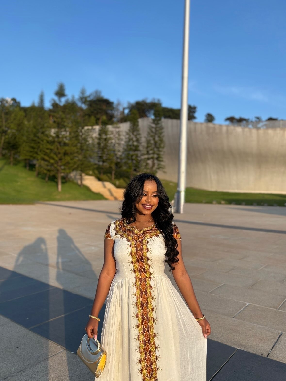 A woman wearing a white Ethiopian clothing with colorful embroidered patterns stands outdoors on a paved plaza with trees and a concrete wall in the background.