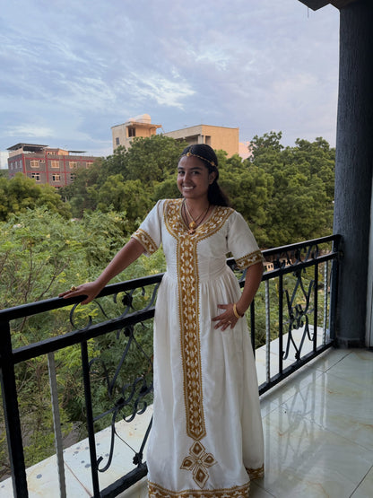 A woman wearing a white traditional embroidered Ethiopia store dress stands on a balcony, holding the railing. The dress features gold embroidery down the front and on the sleeves.