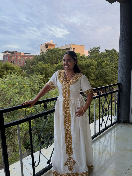 A woman wearing a white traditional embroidered Ethiopia store dress stands on a balcony, holding the railing. The dress features gold embroidery down the front and on the sleeves.