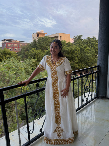 A woman wearing a white traditional Ethiopia store dress with gold embroidery stands on a balcony with a view of trees and buildings in the background.