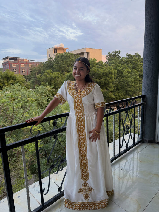 A woman wearing a white traditional Ethiopia store dress with gold embroidery stands on a balcony with a view of trees and buildings in the background.
