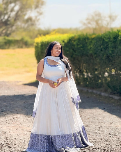 Woman in a white and blue traditional Blue Habesha Zuria standing outdoors with greenery in the background