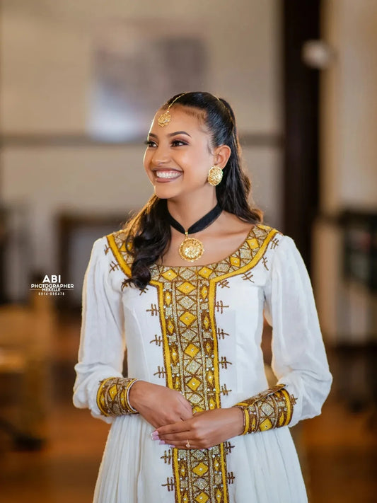 Woman in a white and yellow traditional gold Habesha Libs with jewelry, smiling.
