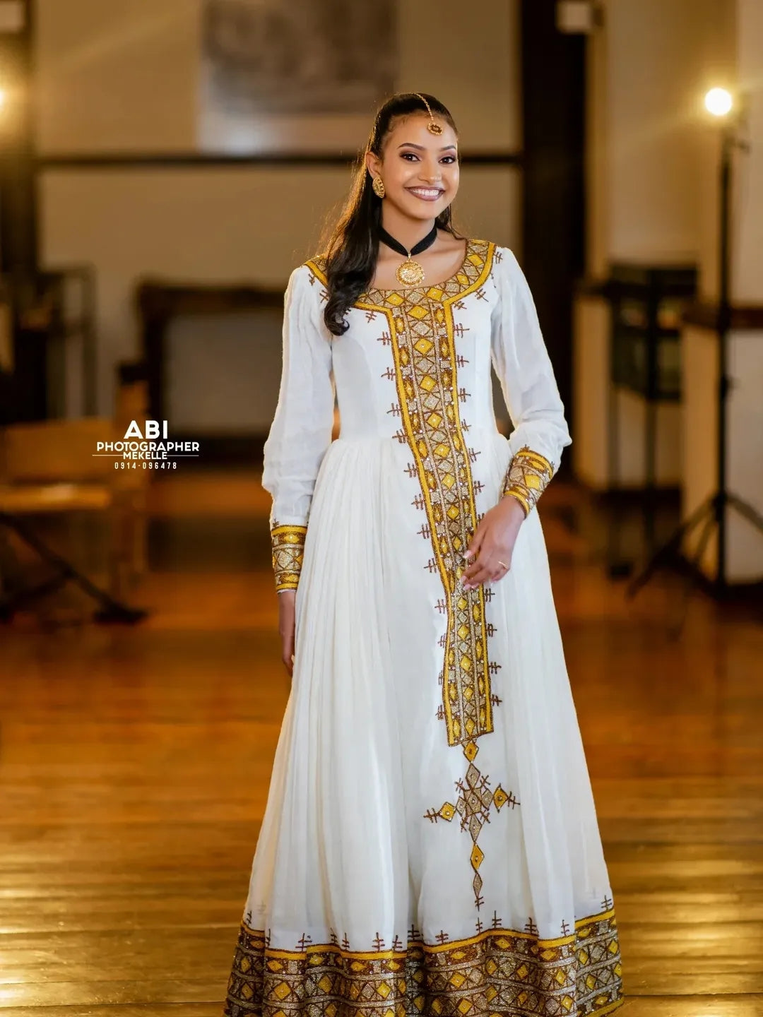 Woman in a white and gold embroidered traditional gold Habesha Libs standing indoors.