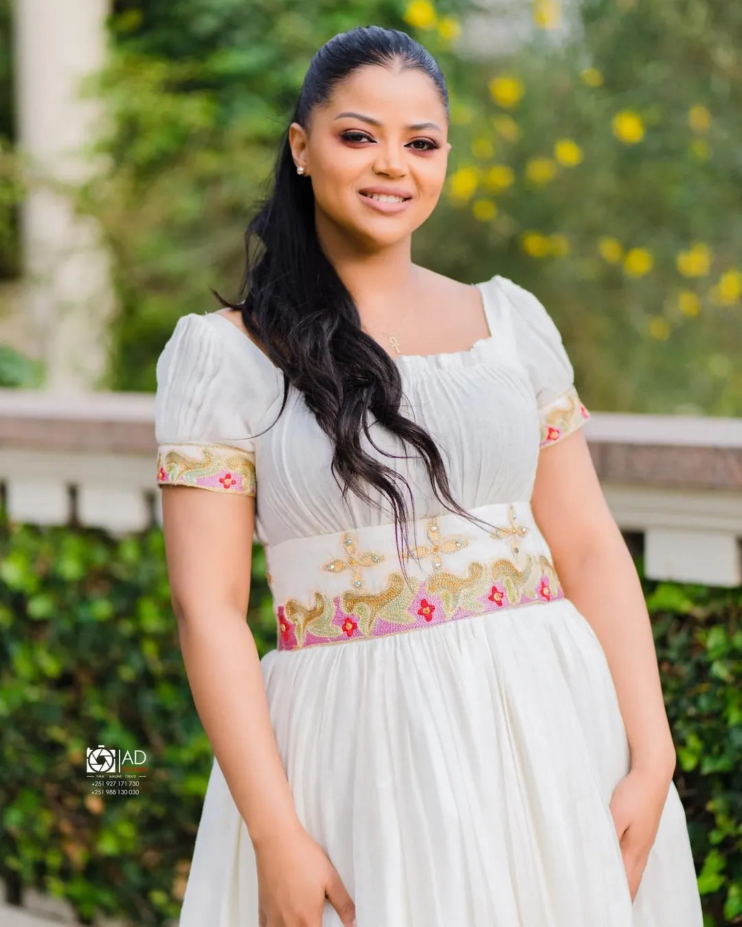 Woman in a white Pink Habesha Zuria with floral embroidery standing outdoors with greenery in the background