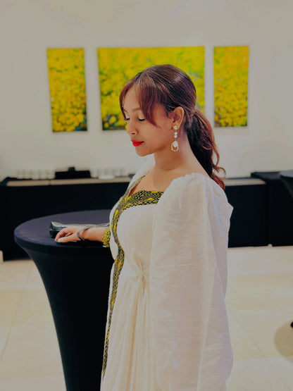 A woman wearing a white long-sleeve Ethiopian shop near me with green and gold embroidery leans on a black table in an indoor setting with yellow floral wall art in the background.